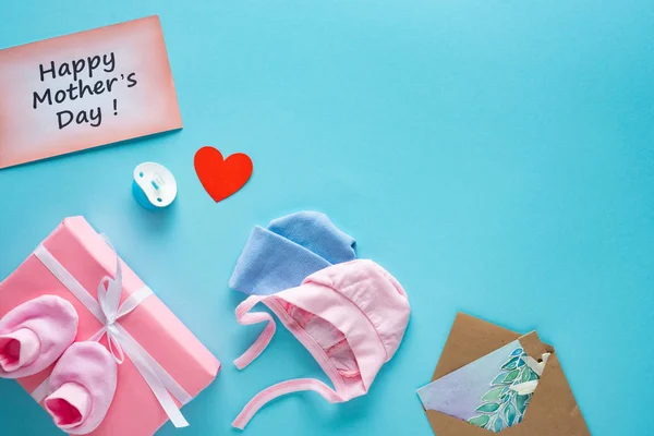 Vista superior de la caja de regalo, ropa de bebé y tarjetas de felicitación con letras felices del día de las madres en la superficie azul - foto de stock