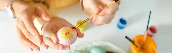 Tiro panorámico de niño pintando huevo de Pascua cerca de la madre en blanco - foto de stock