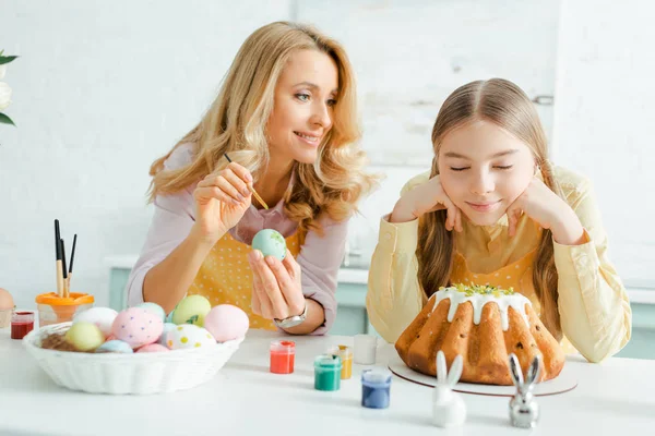 Feliz madre pintando huevo de gallina cerca de hija, conejos decorativos y pastel de Pascua - foto de stock