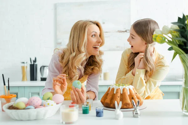Happy money and daughter looking at each other near easter cake, chicken eggs, decorative rabbits and tulips — Stock Photo