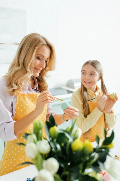 Selective focus of happy daughter and mother painting easter eggs near flowers — Stock Photo