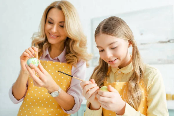 Selective focus of happy daughter and mother painting easter eggs with paintbrushes — Stock Photo
