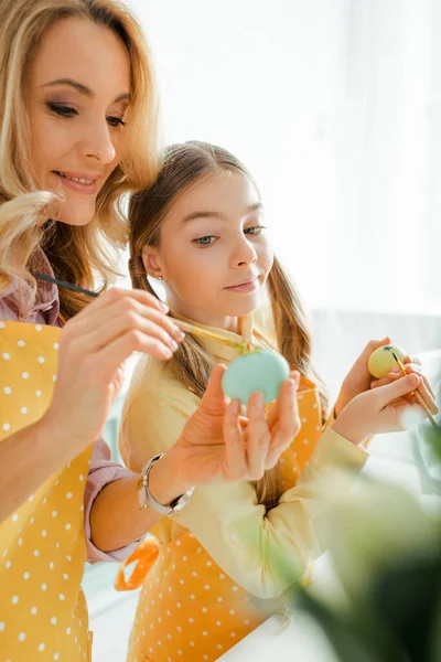 Enfoque selectivo de la hija feliz mirando a la madre pintando huevo de Pascua con pincel - foto de stock