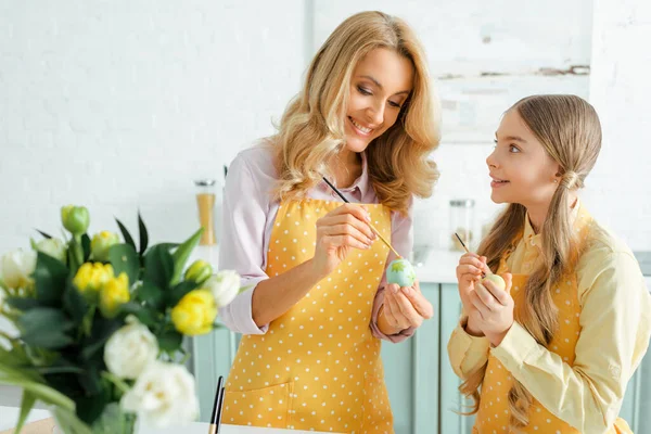 Selektiver Fokus der glücklichen Tochter auf fröhliche Mutter beim Bemalen von Ostereiern mit Pinsel in der Nähe von Tulpen — Stockfoto