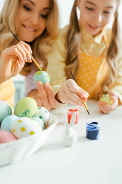 Selective focus of decorative bunny near kid and mother holding paintbrushes while painting easter eggs — Stock Photo