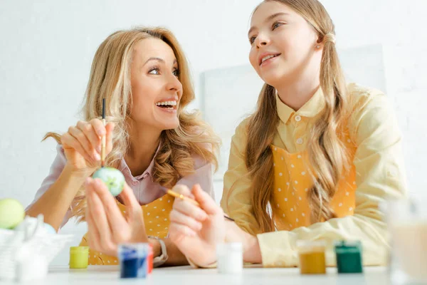 Selective focus of happy mother and daughter holding paintbrushes near jars and easter eggs — Stock Photo