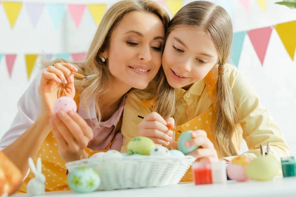 Foco seletivo da filha feliz e mãe pintando ovos de Páscoa perto de coelhos decorativos — Fotografia de Stock