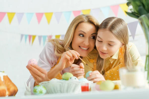 Selective focus of cheerful daughter and mother painting easter eggs at home — Stock Photo