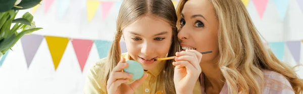 Plano panorámico de la madre sonriendo cerca de hija pintando huevo de Pascua - foto de stock