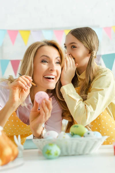 Selektiver Fokus des Kindes flüstert fröhlichen Müttern in der Nähe von Ostereiern ins Ohr — Stockfoto