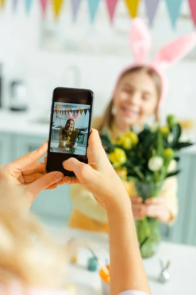 Enfoque selectivo de la madre tomando fotos de linda hija en orejas de conejo con tulipanes - foto de stock