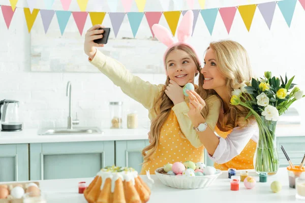 Mãe feliz e filha em orelhas de coelho tomando selfie perto de bolo de Páscoa e ovos de galinha pintados — Fotografia de Stock