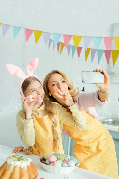Вибірковий фокус щасливої матері і дочки в вухах кролика, що показує знак миру, приймаючи селфі з розфарбованим пасхальним яйцем — стокове фото