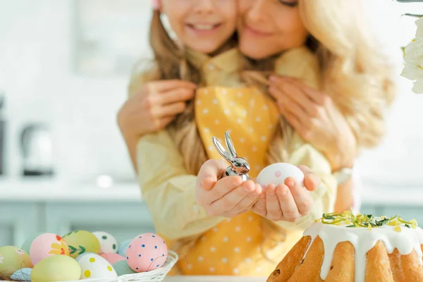 Vista cortada de mãe feliz abraçando filha segurando coelho decorativo e ovo de páscoa pintado — Fotografia de Stock