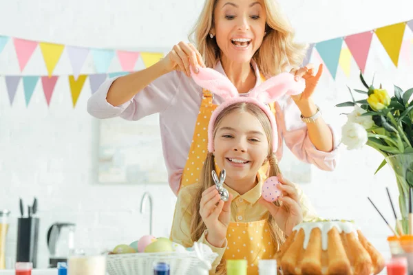 Foco selectivo de madre alegre tocando orejas de conejo en hija feliz sosteniendo conejo decorativo y huevo de Pascua - foto de stock