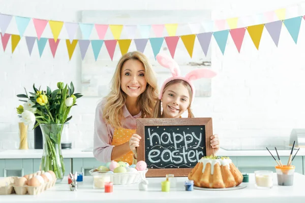 Fröhliche Mutter neben Tulpen und Tochter mit Hasenohren mit Kreidetafel mit fröhlichem Osteraufdruck — Stockfoto