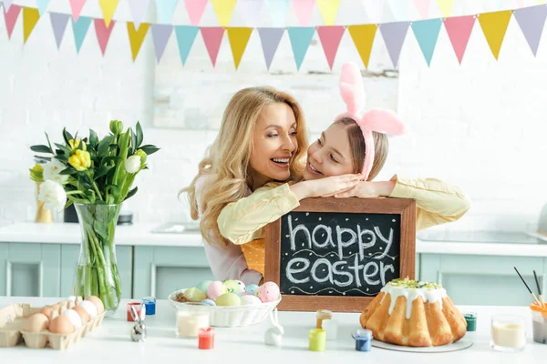 Fröhliche Mutter neben Tochter mit Hasenohren, die Kreidetafel mit fröhlichem Osteraufdruck hält — Stockfoto