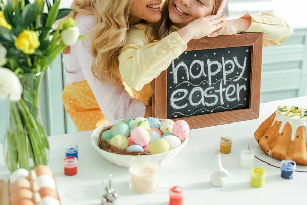 Vista recortada de la madre alegre cerca de la hija sosteniendo pizarra con letras de Pascua feliz - foto de stock