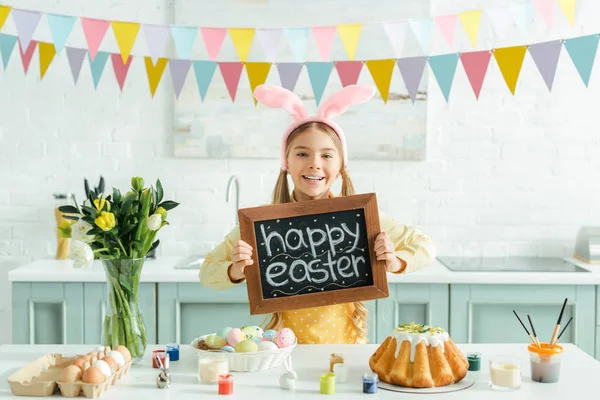 Cheerful kid with bunny ears holding chalkboard with happy easter lettering — Stock Photo