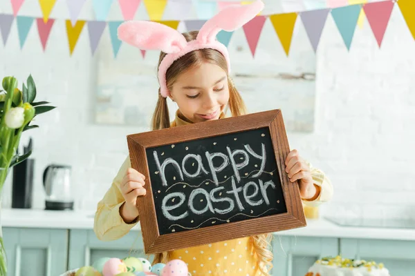 Niño alegre con orejas de conejo mirando pizarra con letras de Pascua feliz - foto de stock