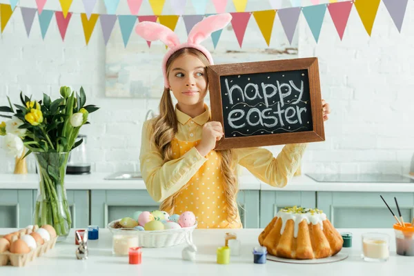 Cute kid with bunny ears holding chalkboard with happy easter lettering and looking away — Stock Photo