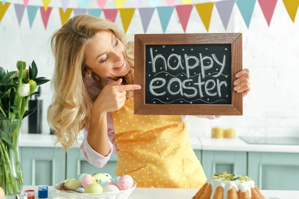 Mujer alegre apuntando con el dedo a la pizarra con letras de Pascua feliz cerca de huevos de pollo pintados - foto de stock