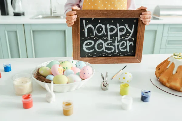 Vista recortada de la mujer sosteniendo pizarra con letras de Pascua feliz cerca de conejos decorativos y huevos de pollo - foto de stock