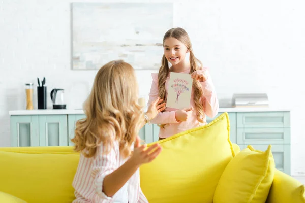 Selective focus of happy kid holding greeting card with happy mothers day lettering near mother — Stock Photo