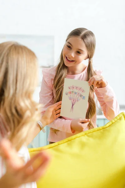 Selective focus of happy daughter holding greeting card with happy mothers day lettering near mother — Stock Photo