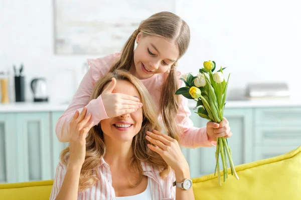 Glückliches Kind bedeckt die Augen der fröhlichen Mutter, während es einen Strauß Tulpen in der Hand hält — Stockfoto