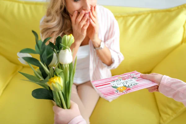 Vista recortada de la madre sorprendida recibiendo tulipanes y tarjeta de felicitación con feliz día de las madres letras de hija - foto de stock
