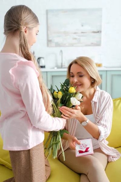 Mãe feliz olhando para tulipas enquanto segurando cartão de saudação perto da filha — Fotografia de Stock