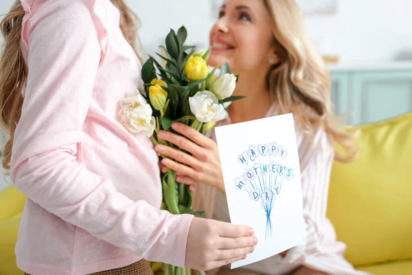 Foco seletivo da criança segurando cartão de saudação com feliz dia das mães lettering e flores perto de mãe feliz — Fotografia de Stock