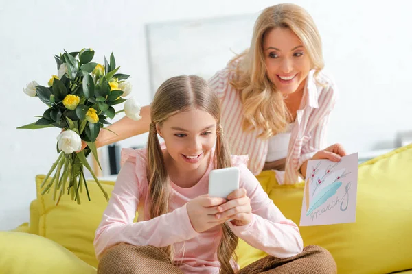 Glückliche Mutter mit Tulpen und Grußkarte mit 8-März-Schriftzug in der Nähe der Tochter per Smartphone — Stockfoto