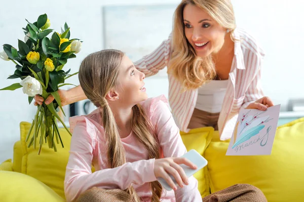 Feliz madre sosteniendo tulipanes y tarjeta de felicitación con 8 letras de marzo cerca de la hija en casa - foto de stock