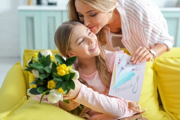 Mãe feliz segurando tulipas e cartão de saudação com 8 letras de marcha enquanto beija a filha em casa — Fotografia de Stock