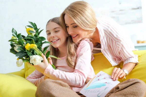 Felice madre che tiene tulipani e biglietto di auguri con 8 lettere di marcia vicino alla figlia sorridente a casa — Foto stock