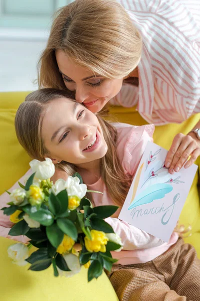 Felice madre in possesso di biglietto di auguri con 8 marzo lettering mentre baciare la figlia a casa — Foto stock