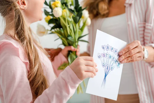 Recortado vista de niño feliz sosteniendo tulipanes y tarjeta de felicitación con feliz día de las madres letras cerca de la madre - foto de stock