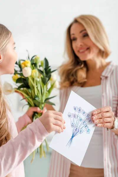 Selective focus of cheerful kid holding tulips and greeting card with happy mothers day lettering near mother — Stock Photo