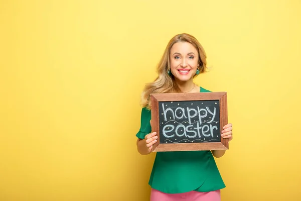 Mujer feliz sosteniendo pizarra con letras de Pascua feliz aislado en amarillo - foto de stock
