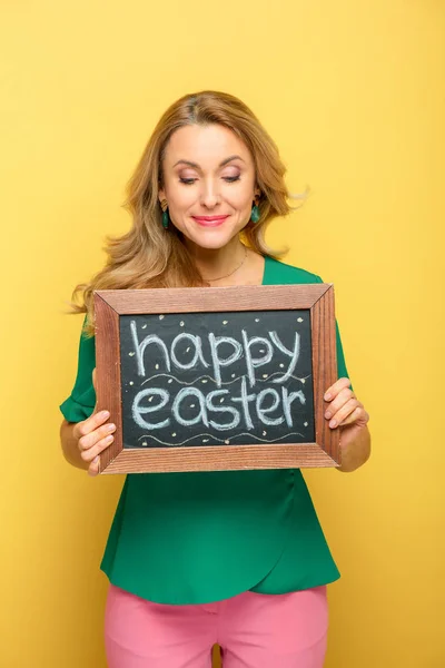 Cheerful woman holding chalkboard with happy easter lettering isolated on yellow — Stock Photo