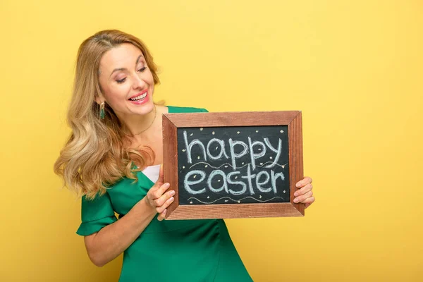 Fröhliche Frau blickt auf Tafel mit fröhlichem Osteraufdruck vereinzelt auf gelb — Stockfoto