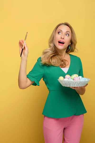 Удивлена женщина, держащая пасхальные яйца и кисть изолированы на желтый — стоковое фото