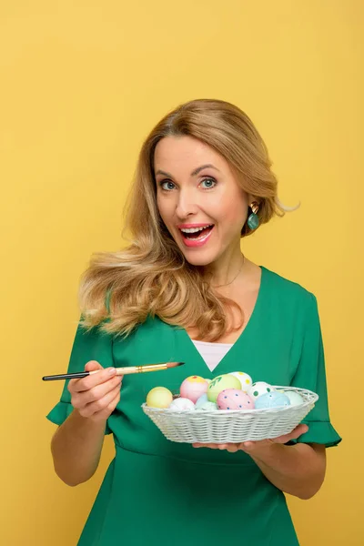 Hermosa mujer sosteniendo pincel cerca de huevos de Pascua aislados en amarillo - foto de stock