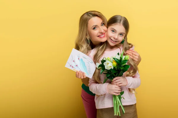 Mãe feliz segurando cartão de saudação com 8 marcha lettering e abraçando filha com tulipas isoladas no amarelo — Fotografia de Stock