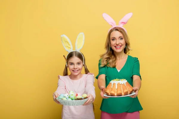 Alegre madre con orejas de conejo sosteniendo pastel de Pascua cerca de hija con huevos de pollo pintados aislados en amarillo - foto de stock