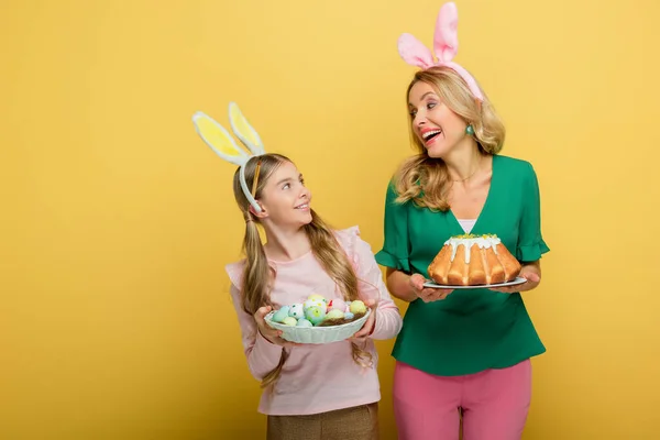 Happy mother with bunny ears holding easter cake near cute daughter with painted chicken eggs isolated on yellow — Stock Photo