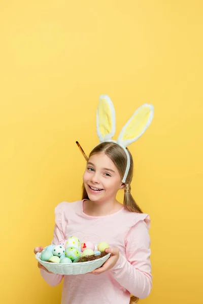 Enfant heureux avec des oreilles de lapin tenant des œufs de Pâques dans le panier isolé sur jaune — Photo de stock