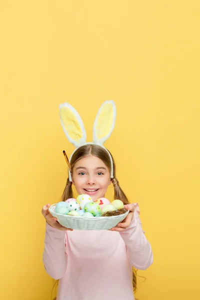 Criança alegre com orelhas de coelho segurando ovos de Páscoa em cesta isolada em amarelo — Fotografia de Stock
